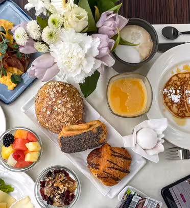 En frokostpålegg med eggerøre og bacon, pannekaker toppet med melis, rundstykker, en croissant, et bakverk, fruktsalat, nøtter, appelsinjuice, kaffe og en smarttelefon. En bukett blomster pynter på bordet.