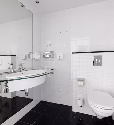 Moderne hvitt bad med vegghengt vask og toalett, stort speil over vasken, og dusjkabinett i glass på høyre side. Sorte gulvfliser står i kontrast til hvite flislagte vegger. Tilbehøret inkluderer såpedispensere og en toalettpapirholder.