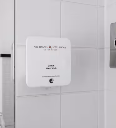 En hvit veggmontert såpedispenser merket "Gentle Hand Wash" er festet til en flislagt baderomsvegg. Til høyre er det montert en krompapirholder med synlige vev. En del av en glass og sort hylle er delvis synlig til venstre.