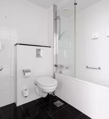 Moderne bad med hvit flislagt vegg og sorte gulvfliser. Funksjonene inkluderer en veggmontert vask, toalett, badekar med glassskjerm og toalettpapirholder. Rommet er lyst og minimalistisk, med et rent og elegant design.