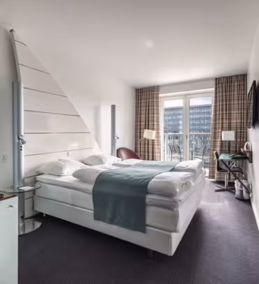 Et moderne hotellrom med stor dobbeltseng, hvitt og blågrønt sengetøy og to høye lamper på hver side. Det er en veggmontert TV, et skrivebord og et stort vindu med gardiner som fører til en balkong. En minibar med drikke er til venstre.