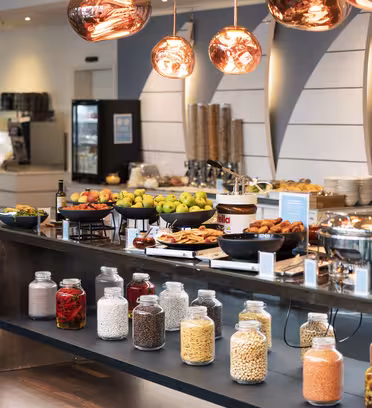 En elegant frokostbuffé med glass med frokostblandinger, nøtter og tørket frukt i forgrunnen. Bakgrunnen har tallerkener med bakverk, brød og frisk frukt, sammen med kobberfargede pendellamper som henger over hodet.