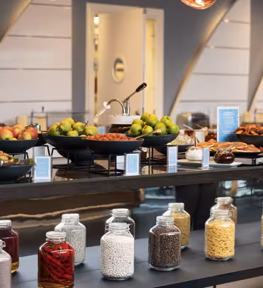 Buffetbord med boller med epler, bakverk og brød. Nedenfor inneholder glasskrukker pasta, bønner og krydder. Bakgrunnen viser et moderne, godt opplyst interiør med geometriske veggdesign og varm belysning.