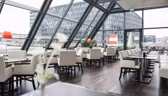 Moderne restaurantinteriør med gulv-til-tak-vinduer, med utsikt over et urbant landskap. Hvite stoler er pent arrangert ved mørke trebord, og små planter gir et snev av grønt. Bygningen utenfor har en rutenettlignende design.