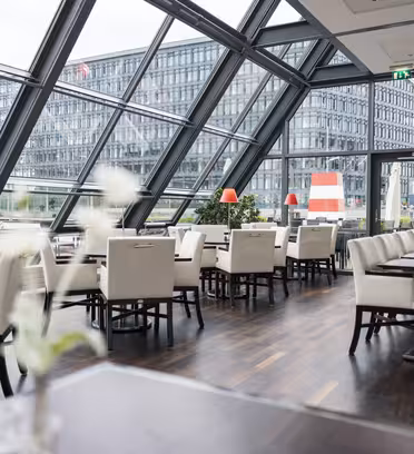 Moderne restaurantinteriør med gulv-til-tak-vinduer, med utsikt over et urbant landskap. Hvite stoler er pent arrangert ved mørke trebord, og små planter gir et snev av grønt. Bygningen utenfor har en rutenettlignende design.
