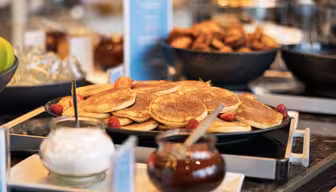 En frokostbuffé med en tallerken med luftige pannekaker pyntet med bringebær. Til dem følger boller med pisket krem og sirup. Bakgrunnen viser flere tallerkener og serveringsredskaper på et trebord.