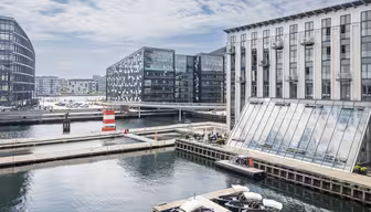 Moderne vannkantsområde med elegante kontorbygg og glassfasader. En bro krysser kanalen, og båter ligger til kai i nærheten. Himmelen er delvis overskyet, og reflekterer over det rolige vannet. En rød og hvit navigasjonsmarkør er synlig i forgrunnen.