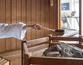En person i en hvit kappe heller vann over varme steiner ved hjelp av en tresleiv i en badstue. Bakgrunnen har trepanel og treningsutstyr synlig gjennom en glassvegg.