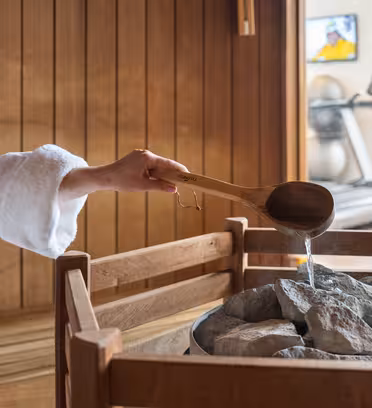 En person i en hvit kappe heller vann fra en tresleiv på varme steiner i et badsturom. Trebenker langs veggene, og et vindu avslører en del av et rom med tredemøller. En TV er montert på veggen.