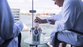 To personer i hvite badekåper sitter på en balkong med utsikt over en vannmasse. En person holder en flaske rosévin over en isbøtte på et glassbord, med vinglass klare til å fylles.