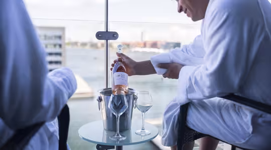 To personer i hvite badekåper sitter på en balkong med utsikt over en vannmasse. En person holder en flaske rosévin over en isbøtte på et glassbord, med vinglass klare til å fylles.