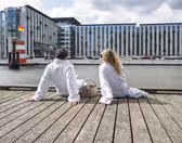 To personer i hvite kapper sitter på en trebrygge, vendt mot en vannmasse. De har en kurv ved siden av seg. I bakgrunnen er moderne bygninger og et lite fyrtårn synlig under en overskyet himmel.