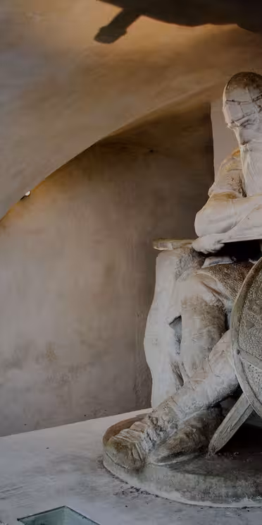 En stor steinstatue av en sittende kriger med langt skjegg og hjelm, hvilende hodet på hånden. Han holder et stort rundt skjold utsmykket med intrikate mønstre. Innstillingen er svakt opplyst, og fremhever statuens detaljer.