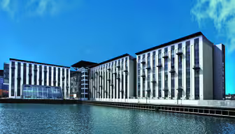 Moderne bygning ved vannkanten under en klar blå himmel, med et sentralt glassatrium og to symmetriske fløyer med balkonger, reflektert i stille vann.