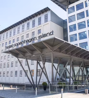 Moderne hotellbygning med baldakininngang støttet av metallbjelker, med store vinduer og et skilt med teksten "Hotel copenhagen island". Bygget er i flere etasjer med en blanding av designelementer i glass og betong.