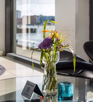 En glassvase med fargerike blomster står på et rundt glassbord ved siden av en blå lysestake. Moderne svarte stoler er i bakgrunnen, med store vinduer som viser utsikt over en by og vann.