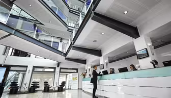 Romslig moderne kontorlobby med stor takhøyde og synlige flere etasjer. En person står ved en elegant resepsjon og snakker med to resepsjonister. Kraftig belysning og glassrekkverk skaper en åpen, moderne atmosfære.