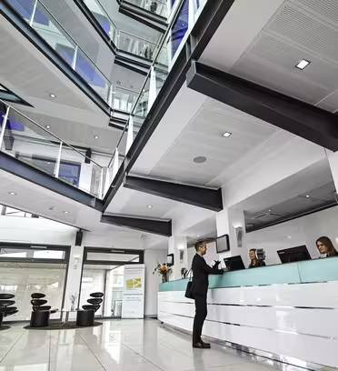Romslig moderne kontorlobby med stor takhøyde og synlige flere etasjer. En person står ved en elegant resepsjon og snakker med to resepsjonister. Kraftig belysning og glassrekkverk skaper en åpen, moderne atmosfære.