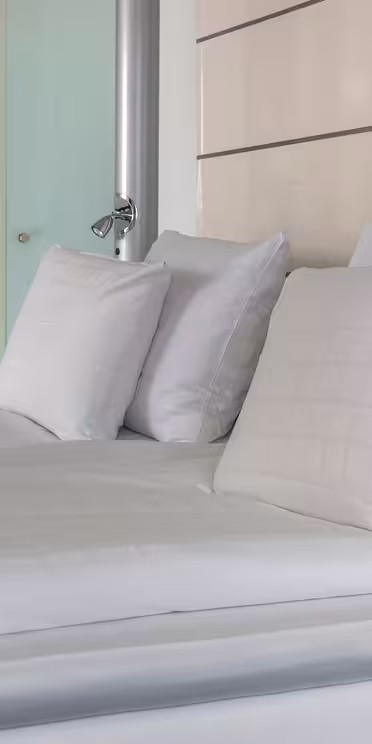 Moderne hotellrom med en pent oppredd seng med hvitt sengetøy og flere puter. En trehylle med drinker, kopper og en vannkoker er i bakgrunnen, ved siden av en frostet glassdør. Rommet har en stilig og minimalistisk design.
