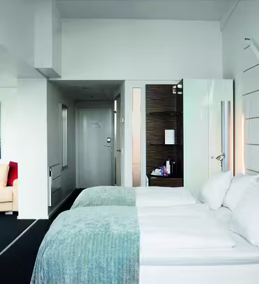Moderne hotellrom med to pent oppredde enkeltsenger, veggmontert TV og kjøkkenkrok med trepanel. En sittegruppe med en beige sofa og røde puter er synlig i bakgrunnen. Lys, minimalistisk innredning forbedrer plassen.