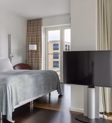 Et moderne hotellrom med en pent oppredd dobbeltseng, flatskjerm-TV på stativ og store vinduer med utsikt over vannkanten og urbane bygninger. Rommet har lyse vegger, tregulv og beige gardiner.