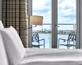 Et stilig hotellrom har en pent oppredd seng med hvitt sengetøy, en lampe og utsikt gjennom glassdører til en industriell vannkant med bygninger og skorsteiner. To stoler sitter på en balkong utenfor, innrammet av stripete gardiner.