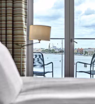 Et stilig hotellrom har en pent oppredd seng med hvitt sengetøy, en lampe og utsikt gjennom glassdører til en industriell vannkant med bygninger og skorsteiner. To stoler sitter på en balkong utenfor, innrammet av stripete gardiner.