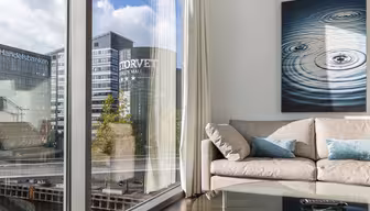 En moderne stue med en beige sofa og blå puter. Et stort vindu gir utsikt over kontorbygg, inkludert Handelsbanken og Torvet Sogn kjøpesenter. Et maleri av krusninger på vann henger på veggen. Et glass salongbord er i forgrunnen.