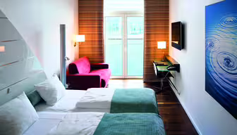 Moderne hotellrom med to pent oppredde enkeltsenger, hvitt og blågrønt sengetøy og et stort bølgemønstret kunstverk på veggen. En rød lenestol og et skrivebord med en lampe er i nærheten av et vindu med brune gardiner med utsikt over en balkong.