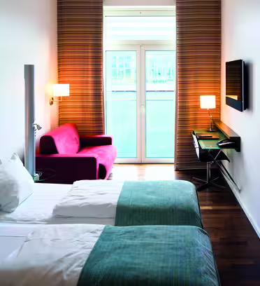Moderne hotellrom med to pent oppredde enkeltsenger, hvitt og blågrønt sengetøy og et stort bølgemønstret kunstverk på veggen. En rød lenestol og et skrivebord med en lampe er i nærheten av et vindu med brune gardiner med utsikt over en balkong.