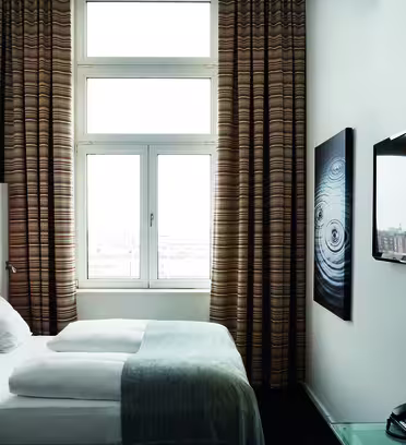 Et moderne hotellrom med en pent oppredd seng, to puter og et lysegråt teppe. Et stort vindu med stripete gardiner viser utsikt over byen. En flatskjerm-TV er montert på veggen overfor sengen, ved siden av abstrakt veggkunst.