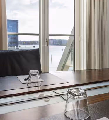 Kontor med trebord og svart skinnstol mot et vindu. Bordet har et omvendt glass og et papir med penn. Vinduet viser utsikt over vannet med bygninger, delvis dekket av hvite gardiner.