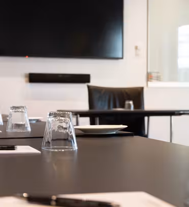 Et konferanserom med stor TV montert på veggen. Et bord er dekket med notatblokker, penner, opp-ned-glass og en tallerken. En svart stol er plassert ved toppen av bordet. Hvite vegger og glasspaneler omgir plassen.
