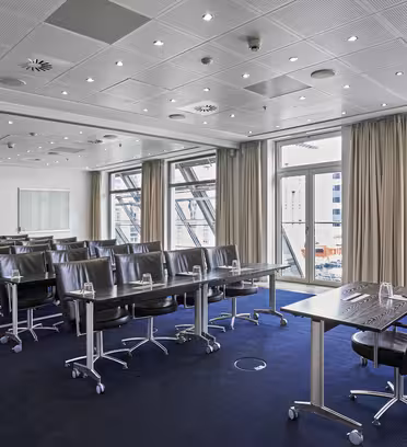 Moderne konferanserom med rader med bord og svarte stoler mot et langt presentasjonsbord. Store vinduer med beige gardiner gir utsikt over en bygning. Rommet har hvitt tak med spotlights og mørkeblått teppegulv.