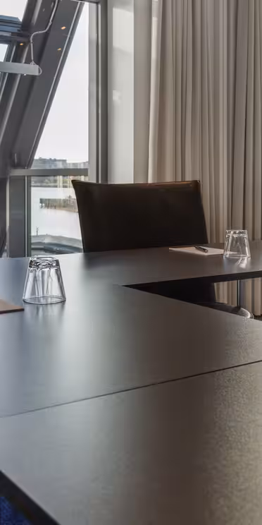 Et moderne konferanserom med store vinduer og utsikt over en elv. Sorte bord er arrangert i en U-form, hver med en notisblokk, penn og opp-ned briller. Kremfargede gardiner rammer inn vinduene, og forsterker rommets elegante utseende.