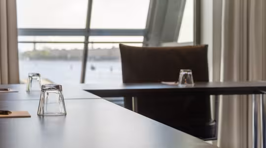 Nærbilde av et konferanseromoppsett med tomme glass snudd på bordene. Store vinduer gir en uskarp utsikt over en vannmasse og fjerntliggende landskap. Rommet er dempet opplyst, og skaper en rolig atmosfære.