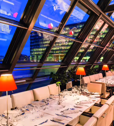 En moderne restaurant med store skrå glassvinduer med utsikt over byens bygninger over en elv. Bordene er dekket med hvite duker, glass og oransje lamper. Scenen er opplyst med varmt lys, noe som skaper en koselig atmosfære.