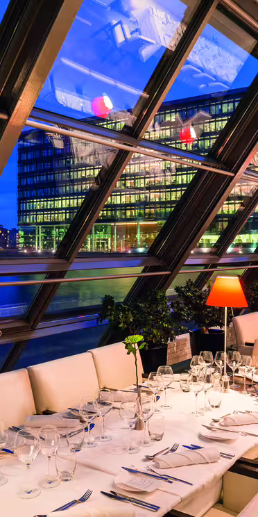 En moderne restaurant med store skrå glassvinduer med utsikt over byens bygninger over en elv. Bordene er dekket med hvite duker, glass og oransje lamper. Scenen er opplyst med varmt lys, noe som skaper en koselig atmosfære.