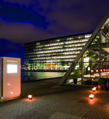Moderne bygg med en trekantet glassfasade opplyst om natten, ved siden av vannreflekterende lys. Et tent skilt viser "Havnen" nær inngangen. Små flammer langs stien, gir varme til scenen under en overskyet himmel.