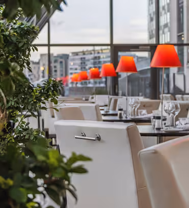 Moderne restaurant med hvite stoler og bord, pent dekket med glass og bestikk. Knallrøde lamper gir mykt lys. Store vinduer gir utsikt over urbane bygninger. Grønn bladplante i forgrunnen gir et preg av natur.