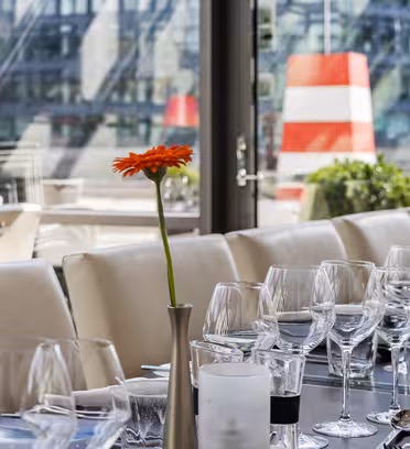 Et moderne spisebord i en restaurant med glassvegger. Hvite stoler omgir bordet, pyntet med klare vinglass, servietter og enkelt bestikk. Lyse oransje gerbera tusenfryd i vaser gir en nyanse av farger til det elegante bordarrangementet.