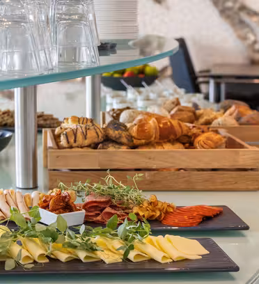 En buffé med skivede oster, kjøtt og ferske grønnsaker på et svart fat i forgrunnen. Bak er brett med diverse brød og bakverk. Klare glass er stablet opp ned på en glasshylle over. Et bord og stoler er i bakgrunnen.