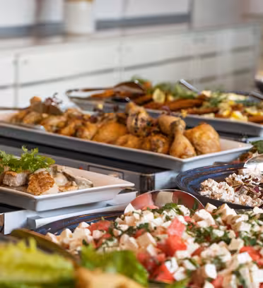 En buffet med en rekke retter, inkludert brett med stekte kyllinglår, blandet salat med feta og vannmelon, pasta og garnerte retter med friske grønnsaker. Tallerkener er pent arrangert på en benkeplate.