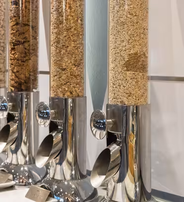 Nærbilde av en rad med kornblandingsdispensere i skinnende metall fylt med forskjellige typer frokostblandinger. Hver dispenser har en spak i bunnen for dispensering. Dispenserne er pent justert mot en flislagt vegg på en disk.