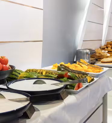 Et buffébord med skåler med oppskåret frukt, hele epler og brett med brød og bakverk. To store skåler med yoghurt er i forgrunnen. Bordet er dekket i et godt opplyst rom med moderne, elegant design.