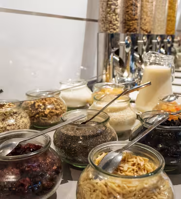 Glasskrukker fylt med forskjellige frokostpålegg som nøtter, tørket frukt og frø står på rekke og rad på en disk, med en beholder med melke- og frokostblandinger i bakgrunnen.