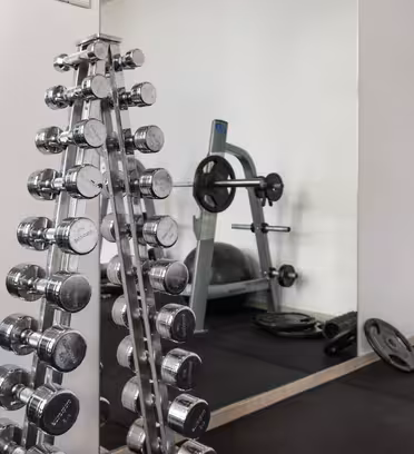 Et treningsstudio med et stativ med skinnende manualer til venstre, en vektbenk med vektstang i bakgrunnen, og ulike vektskiver spredt på gummigulvet. Et stort speil reflekterer utstyret, og forsterker den romslige følelsen.